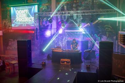 Deux DJ mixent sur une scène éclairée par des projecteurs colorés, devant un écran affichant ’Don't Care Project’.