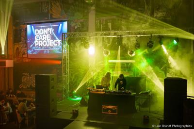 Deux DJ mixent sur une scène éclairée par des projecteurs colorés, devant un écran affichant ’Don't Care Project’.