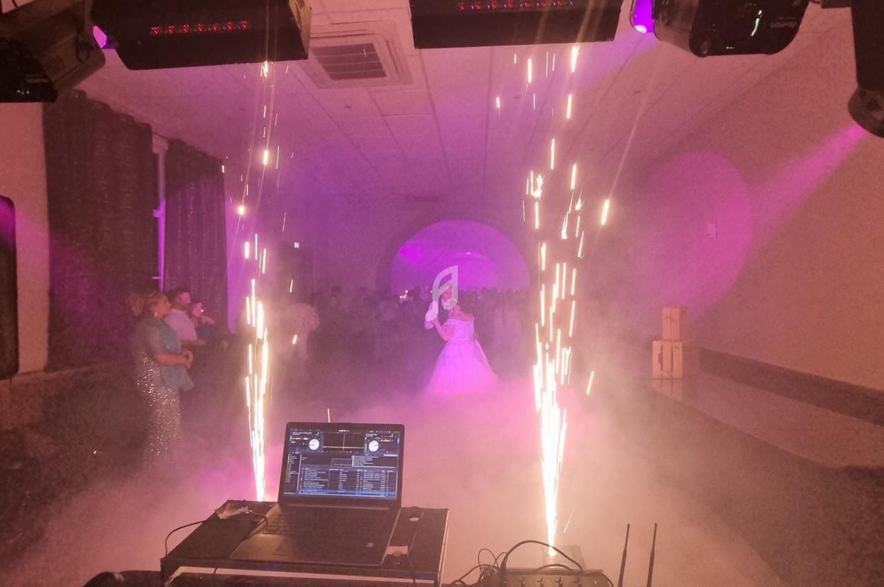 Salle de réception avec éclairage violet, fumée, étincelles et une mariée dansant au centre, vue depuis une console DJ.