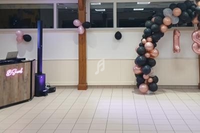 Décoration de fête avec arche de ballons noirs, gris et roses, et une table de DJ équipée de matériel audio.
