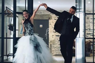 Un couple en tenue de mariage entre dans une pièce lumineuse avec des murs en pierre et un sol en bois.