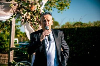 Un homme tenant un micro parle en extérieur, sous une arche décorée de fleurs, par une journée ensoleillée.