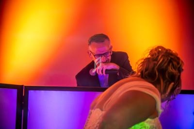 Un DJ souriant derrière sa table de mixage, éclairé par des lumières colorées, avec une personne dansant au premier plan.