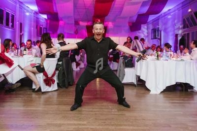 Un homme souriant, micro en main, pose de manière expressive dans une salle de réception animée.