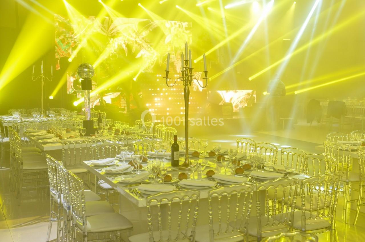 Salle de réception illuminée par des projecteurs jaunes, avec tables dressées et chaises transparentes.