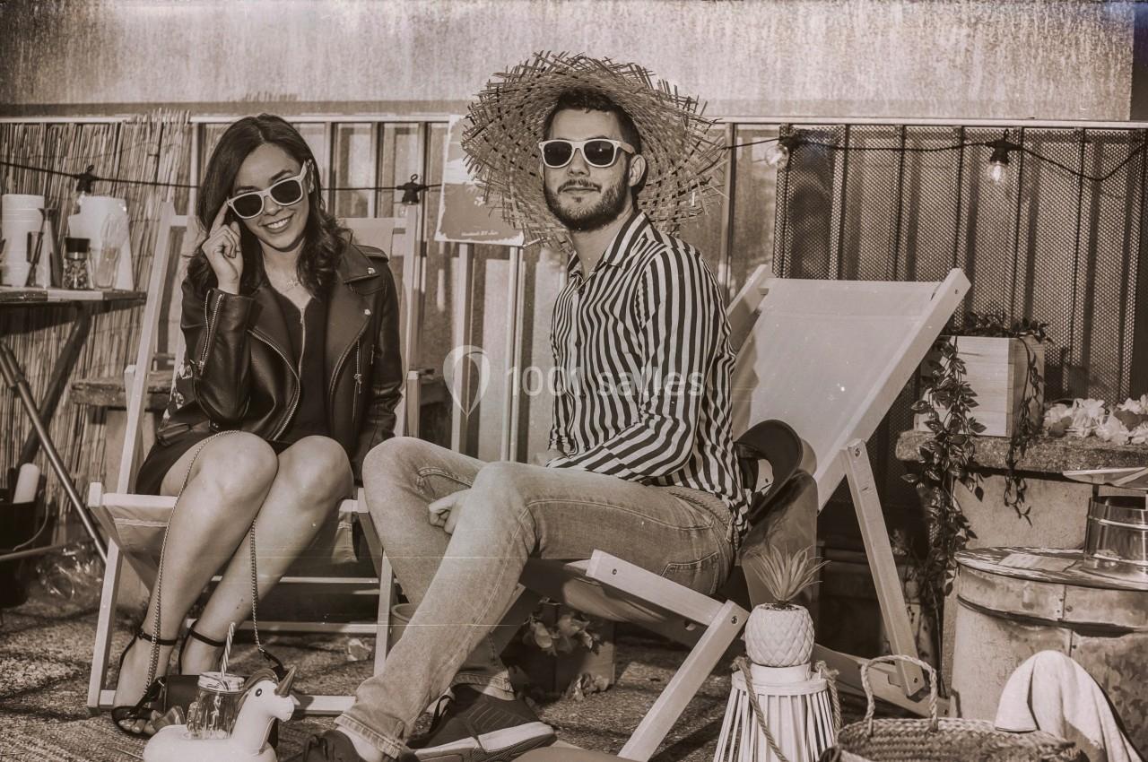 Un homme et une femme assis sur des chaises longues, portant des lunettes de soleil, dans un décor rétro en extérieur.