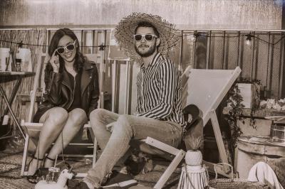 Un homme et une femme assis sur des chaises longues, portant des lunettes de soleil, dans un décor rétro en extérieur.