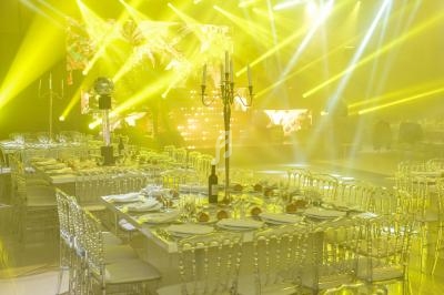 Salle de réception illuminée par des projecteurs jaunes, avec des tables dressées et des chaises transparentes.