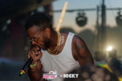 Un chanteur en débardeur blanc et lunettes de soleil interprète sur scène avec un micro, éclairé par des projecteurs.