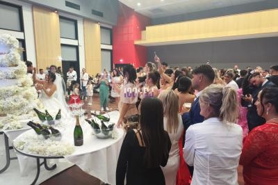 Groupe de personnes rassemblées dans une salle de réception, observant un couple près d'un grand gâteau de mariage.