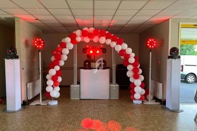 Arche de ballons rouges et blancs entourant une table de DJ éclairée par des projecteurs dans une salle de réception.