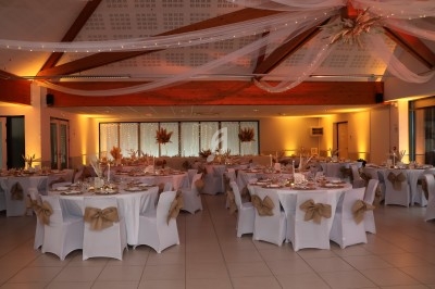 Salle de réception décorée avec des tables rondes, nappes blanches, chaises ornées de nœuds beige et éclairage chaleureux.