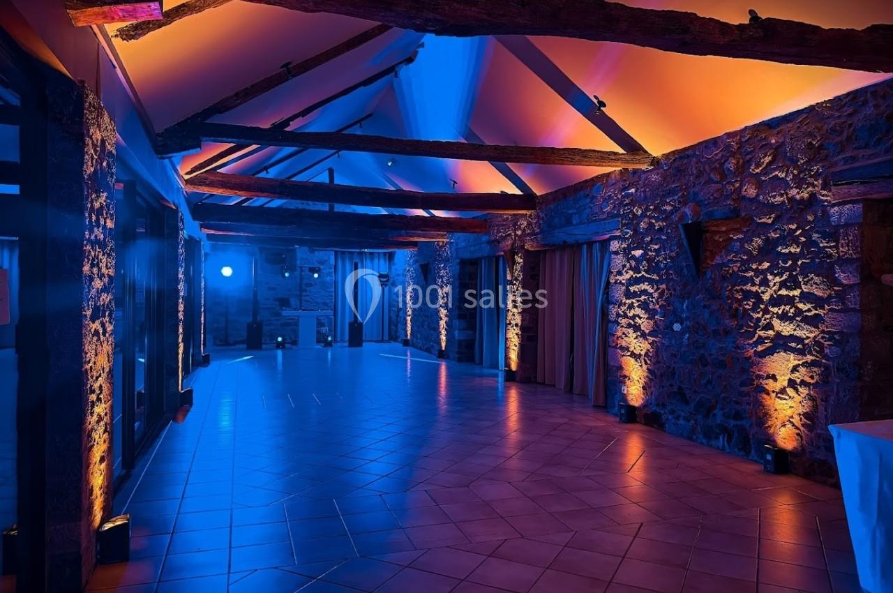 Salle en pierre avec poutres apparentes, éclairée par des lumières colorées bleues et orange, au plafond incliné.