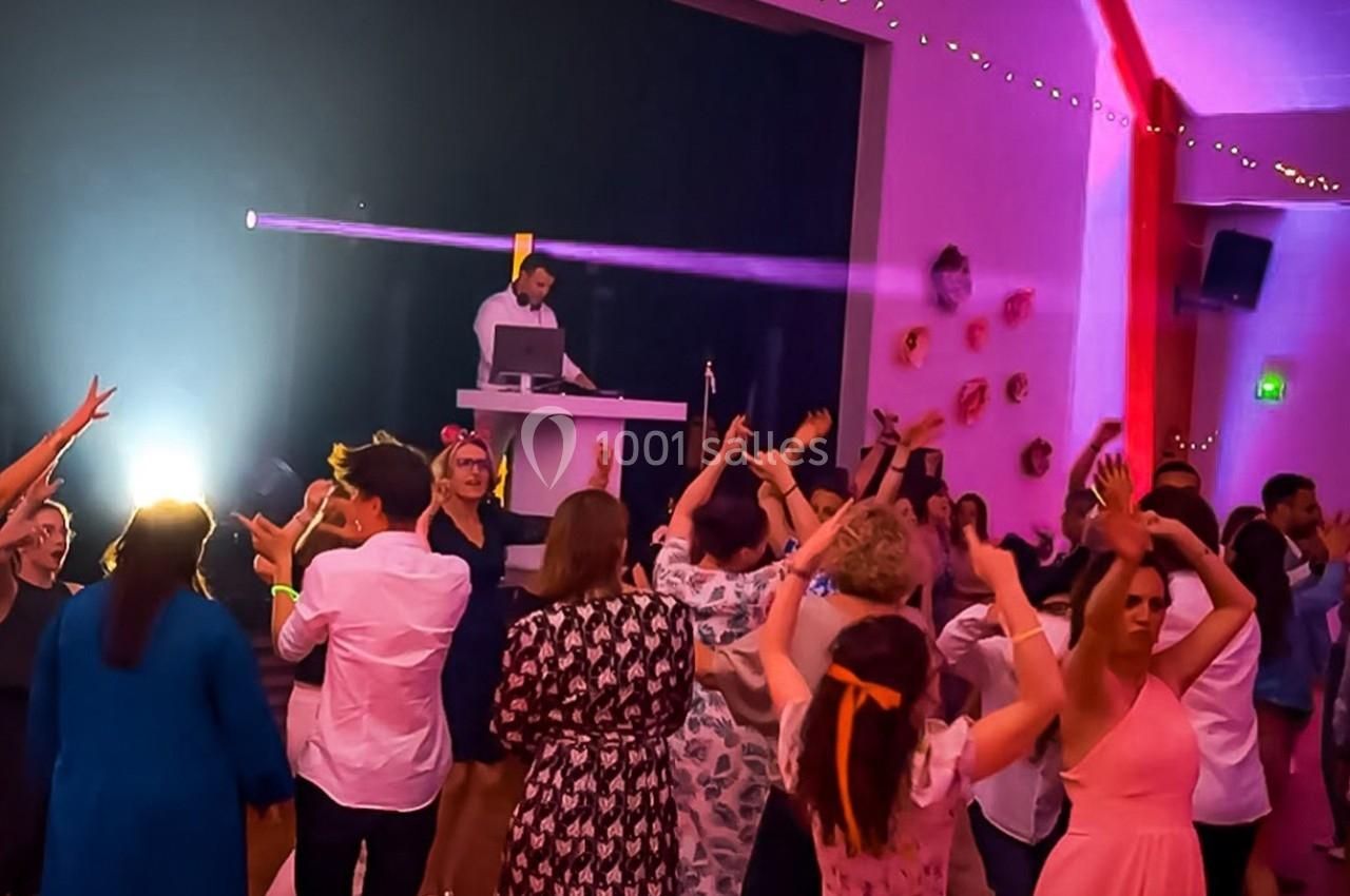 Des personnes dansent dans une salle éclairée par des lumières colorées, avec un DJ en arrière-plan sur une estrade.