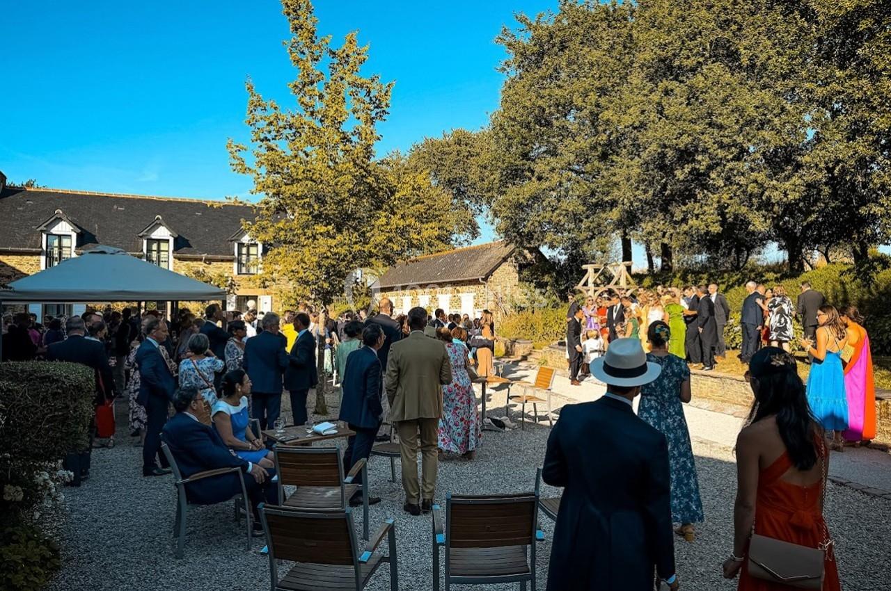 Groupe de personnes rassemblées en extérieur lors d'un événement, près de bâtiments en pierre et d'arbres sous un ciel bleu.