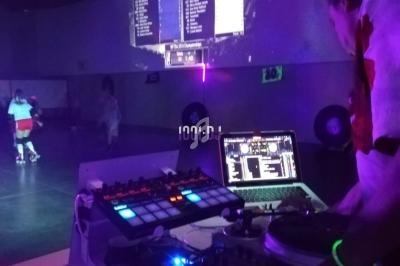 Un DJ mixe sur une table de mixage dans une salle sombre avec des lumières violettes et des patineurs en arrière-plan.