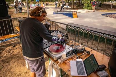 Un DJ mixe sur une platine vinyle sous une tente, près d'un skatepark en plein air.