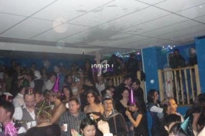 Salle bondée avec des personnes célébrant, portant des chapeaux festifs et tenant des boissons, dans une ambiance animée.