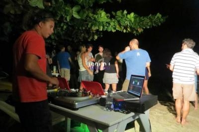 Un DJ mixe sur une plage de nuit tandis que des personnes dansent sous des arbres.