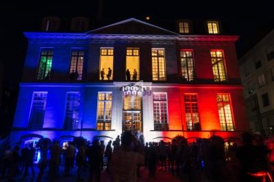 Façade d'un bâtiment illuminé en bleu, blanc et rouge, avec des silhouettes de personnes rassemblées devant.