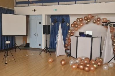 Salle décorée pour un événement avec arche de ballons dorés et blancs, écran de projection et matériel audio.