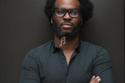 Homme portant des lunettes et une chemise à motifs, debout devant un fond sombre, les bras croisés.