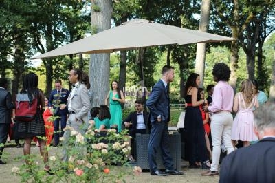 Personnes discutant en plein air sous un grand parasol dans un jardin, entourées de fleurs et d'arbres.