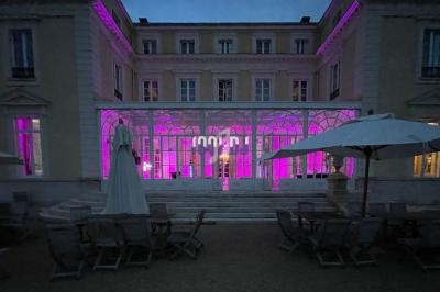 Façade d'un bâtiment éclairé de lumières roses, avec une terrasse équipée de parasols et de chaises en soirée.