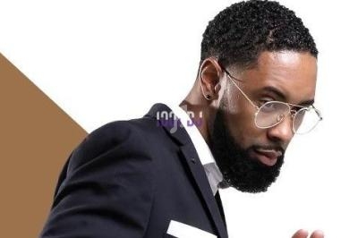 Homme barbu portant des lunettes et un costume sombre, regardant sur le côté, sur fond blanc et marron.