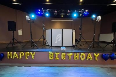 Scène décorée pour un anniversaire avec ballons ’Happy Birthday’, éclairages colorés et matériel de sonorisation.