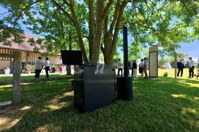 Équipement audio installé en extérieur sous un arbre, avec des personnes rassemblées en arrière-plan.