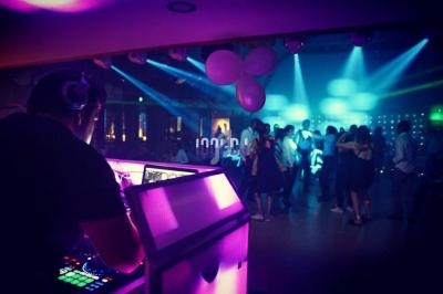 Un DJ mixe sur une scène éclairée en violet tandis qu'un groupe de personnes danse sous des lumières colorées.