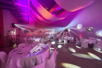 Salle de réception décorée avec des tables rondes, nappes blanches, éclairages colorés et voilages suspendus.