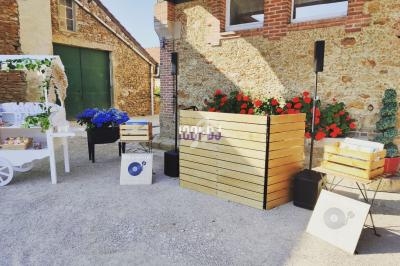 Stand de DJ en extérieur avec des caisses en bois, des fleurs rouges et bleues, et un chariot décoratif blanc.