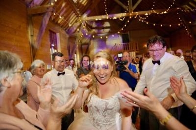 Une mariée souriante danse entourée d'invités applaudissant dans une salle en bois décorée de guirlandes lumineuses.