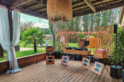 Espace extérieur couvert avec platines DJ, décor tropical, plantes, rideaux blancs et panneaux en bois.