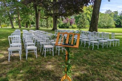 Chaises blanches alignées en extérieur sous des arbres, avec un pupitre en bois décoré de feuillage au premier plan.