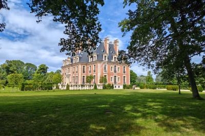 Manoir en briques rouges entouré d'un parc verdoyant, avec des arbres et un ciel partiellement nuageux.