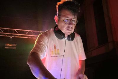 Un DJ mixe sur une table de mixage éclairée, dans une ambiance sombre avec des lumières colorées en arrière-plan.