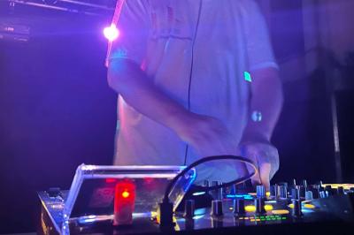 Un DJ mixe sur une table de mixage éclairée, dans une ambiance sombre avec des lumières colorées en arrière-plan.