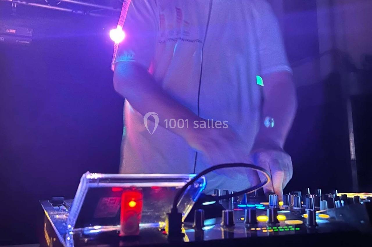 Un DJ mixe sur une table de mixage éclairée, dans une ambiance sombre avec des lumières colorées en arrière-plan.