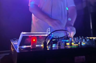Un DJ manipule une table de mixage éclairée par des lumières colorées dans une ambiance sombre.