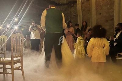 Des personnes dansent et discutent dans une salle faiblement éclairée, avec de la fumée au sol et des lumières en arrière…