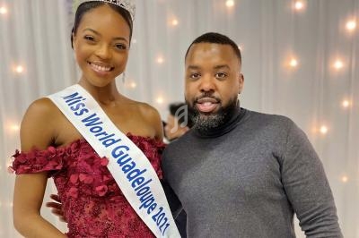 Une femme portant une écharpe ’Miss World Guadeloupe 2021’ pose avec un homme devant un fond lumineux.