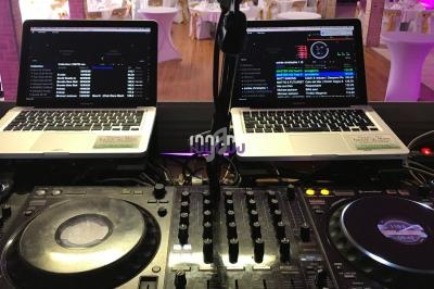 Poste de DJ avec deux ordinateurs portables affichant des playlists, platines et table de mixage en arrière-plan d'une salle…