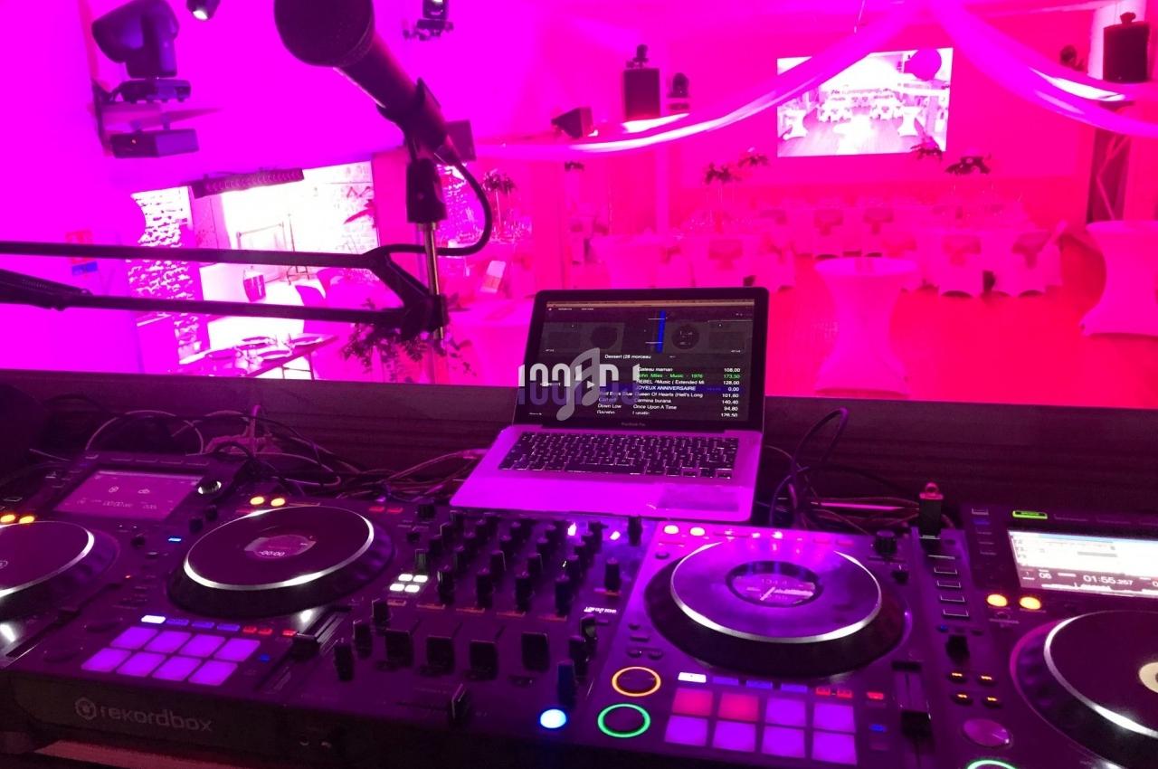 Poste de DJ avec platines, table de mixage et ordinateur, dans une salle décorée de lumières roses pour un événement.