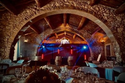 Salle voûtée en pierre avec tables dressées, éclairages colorés et barils décoratifs au fond.