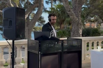 Un DJ en costume mixant de la musique en extérieur, sur une terrasse entourée d'arbres et avec vue sur la mer.