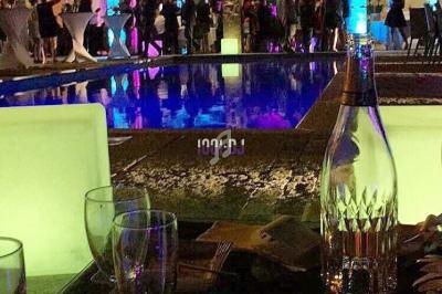 Vue nocturne d'une fête près d'une piscine éclairée, avec des tables dressées et des invités en arrière-plan.