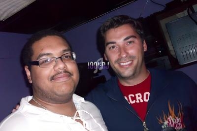 Deux hommes posant côte à côte dans une pièce sombre avec des équipements électroniques en arrière-plan.